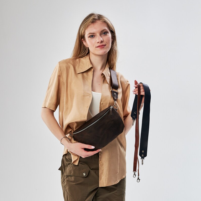 Puede incluir: Una ri&ntilde;onera de ante marr&oacute;n con cierre de cremallera y hebilla plateada. La bolsa la lleva una persona que lleva una camisa beige y pantalones cargo verdes. La bolsa est&aacute; unida a una correa marr&oacute;n y negra.
