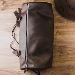 Puede incluir: Una bolsa de viaje de cuero marr&oacute;n con cierre de cremallera y una correa. La bolsa est&aacute; sobre una superficie de madera.