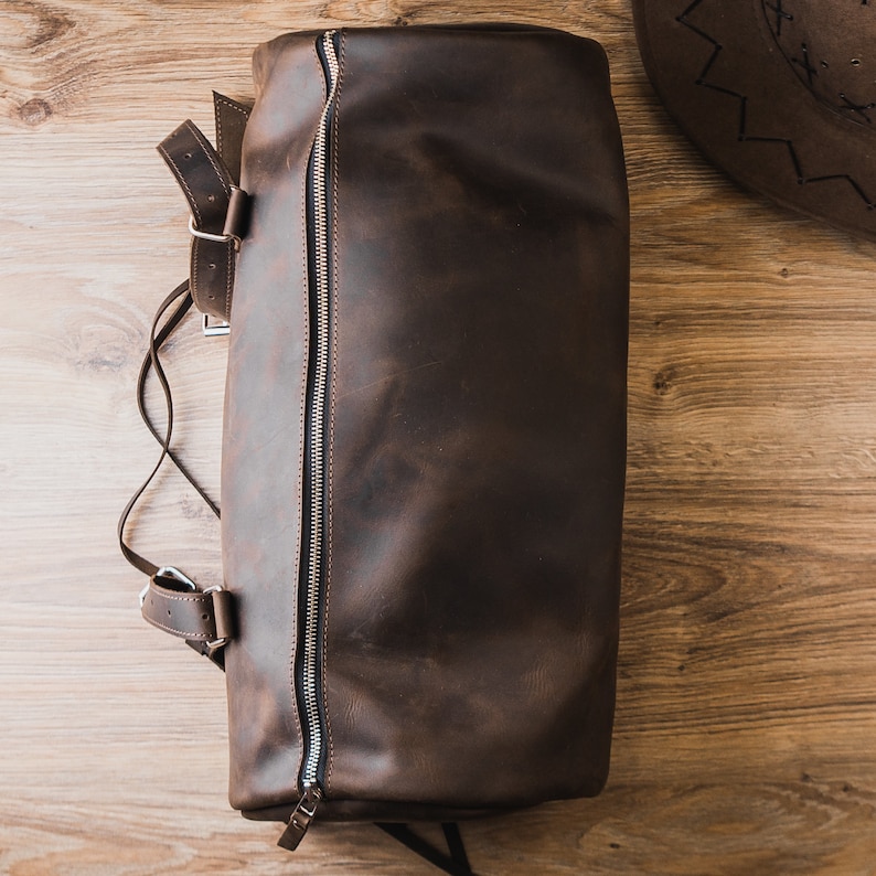 Puede incluir: Una bolsa de viaje de cuero marr&oacute;n con cierre de cremallera y una correa. La bolsa est&aacute; sobre una superficie de madera.