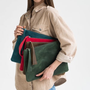May include: A set of four leather clutches in different colors: teal, red, olive green, and dark green. The clutches are stacked on top of each other and held by a person.