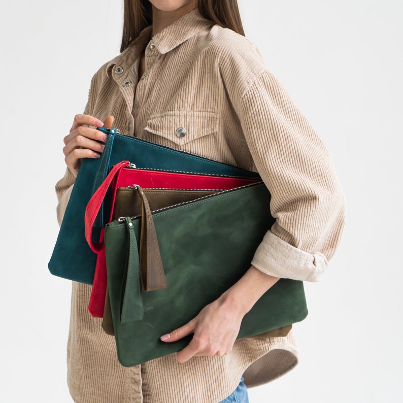 May include: A set of four leather clutches in different colors: teal, red, olive green, and dark green. The clutches are stacked on top of each other and held by a person.
