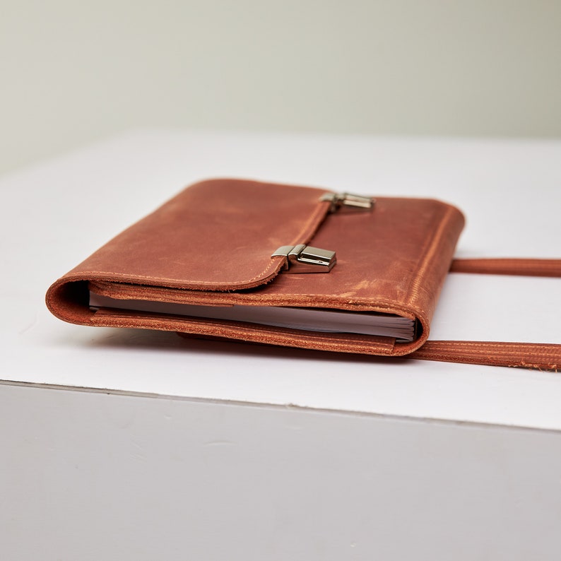 May include: A brown leather journal with a flap closure and silver clasps. The journal is closed and lying on a white surface.