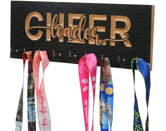 May include: A black wooden medal display with the word "CHEER" carved into it in a light brown colour. Below the word are several hooks holding colourful ribbons. The ribbons are pink, blue, and patterned.