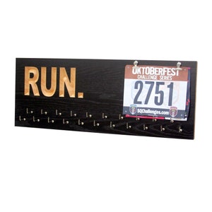 May include: A black wooden medal display with the word "RUN." in gold lettering. The display has 20 hooks for hanging medals and a race bib with the number "2751" attached to the top.