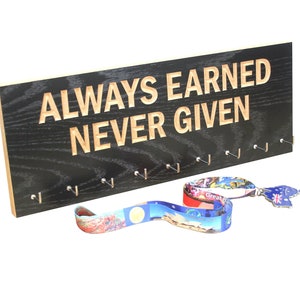 May include: Black wooden medal display with the text "ALWAYS EARNED NEVER GIVEN" engraved in a light wood color. The display has 10 hooks for hanging medals. Two colorful medal ribbons are hanging from the hooks.