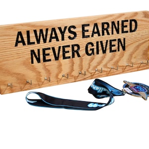 May include: A wooden medal display rack with the text "ALWAYS EARNED NEVER GIVEN" in black lettering. The rack has 12 hooks for hanging medals.