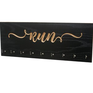 May include: A black wooden wall-mounted display with the word "RUN" carved in a light wood tone. Below the word are eight small metal hooks. The display is designed to hold medals or other items.
