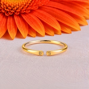 Op de afbeelding: Een goudkleurige open ring met een dunne, gepolijste band. De twee uiteinden van de ring zijn afgeplat en bevatten elk een gegraveerde letter, met de letter B aan de linkerkant en de letter S aan de rechterkant van de opening.