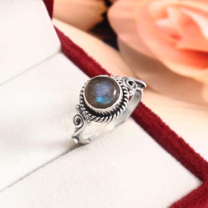 May include: A silver ring featuring a round, iridescent labradorite gemstone set in a twisted rope-style bezel. The band is crafted with decorative scrollwork details on the sides, framing the central stone with a polished metallic finish.
