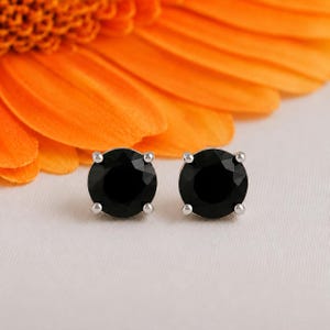 Puede incluir: Un par de pendientes de botón redondos con piedras negras facetadas engastadas en garras de metal plateado. Cada pendiente tiene una silueta circular con cuatro pequeñas garras que aseguran la piedra oscura y reflectante en su lugar.