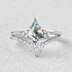 May include: A two-piece ring set featuring a silver band and a matching curved band. The primary ring showcases a kite-shaped moss agate stone with green and white patterns, surrounded by small, clear accent stones.