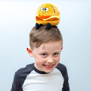 May include: A young boy with freckles smiles at the camera while wearing a plush hamburger toy on his head. The hamburger is yellow and brown with a red tomato and a white sesame seed bun. The toy has the words "In a Million" printed on the side.