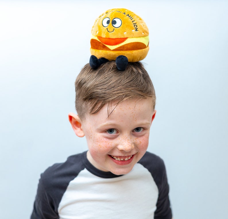 May include: A young boy with freckles smiles at the camera while wearing a plush hamburger toy on his head. The hamburger is yellow and brown with a red tomato and a white sesame seed bun. The toy has the words "In a Million" printed on the side.