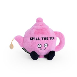 May include: A pink plush teapot with the phrase "SPILL THE TEA" in black text. The teapot has a smiling face, black feet, and a beige tassel. The brand name "munchkins" is on a black tag.