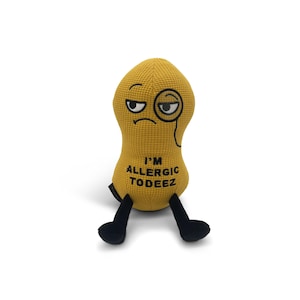 May include: A plush peanut toy with a yellow body, black legs, and a grumpy face with a monocle. The text "I'M ALLERGIC TODEZ" is embroidered on the front. The toy is designed to sit upright.