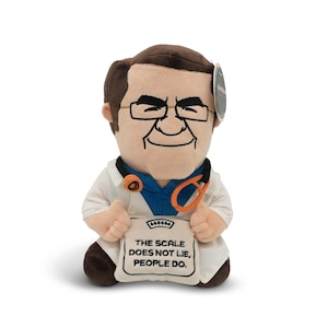 May include: A plush doll of a doctor wearing glasses, a white coat, and a blue shirt. The doll holds a sign that reads, "THE SCALE DOES NOT LIE, PEOPLE DO." The doll has brown hair and a stethoscope around its neck.