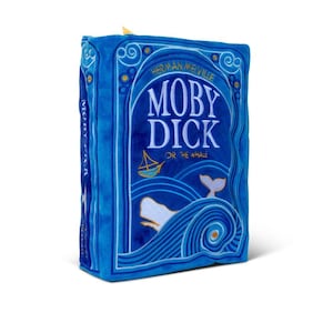 May include: A blue plush book shaped like the cover of Moby Dick by Herman Melville. The cover features a white whale with a blue wave and a small boat in the background. The title "Moby Dick" is in white text with the subtitle "Or The Whale" in smaller white text.