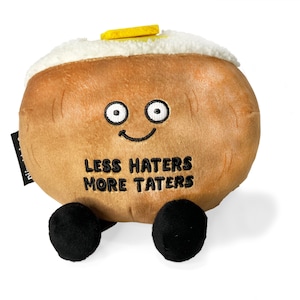 May include: A brown plush potato with a smiling face, wearing a white fuzzy hat with a yellow butter pat on top. The potato has black eyes and a black mouth with the text "Less Haters More Taters" embroidered on the front. The potato has two black plush legs.