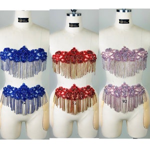 Puede incluir: Tres adornos de cuerpo con cuentas y flecos, uno azul, uno rojo y uno morado, expuestos en maniquíes. Cada pieza presenta una parte superior con cuentas y una parte inferior con cuentas con flecos.