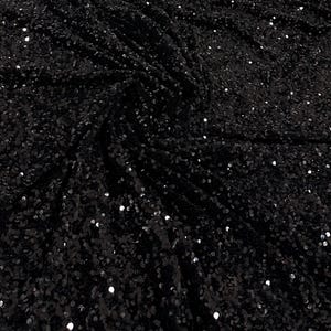 Può includere: Primo piano di tessuto nero ricoperto di piccole paillettes riflettenti. Il tessuto è drappeggiato, creando ombre e riflessi. Le paillettes catturano la luce, conferendo al materiale un aspetto scintillante. L'effetto complessivo è di lusso e glamour.