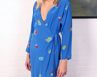 Pode incluir: Um vestido comprido de manga comprida, estilo envelope, em azul vibrante. O vestido apresenta um decote em V profundo e é adornado com formas abstratas coloridas em vermelho, amarelo e azul. O vestido é amarrado na cintura.
