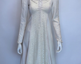 Puede incluir: Vestido crema de manga larga sobre un maniquí. El vestido presenta un corpiño con cordones, un panel de encaje decorativo en la parte delantera y ribete de encaje a lo largo de las mangas y el dobladillo. El vestido tiene un escote redondo y una falda fluida.
