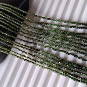 Puede incluir: Se muestran hebras de pequeñas cuentas verdes facetadas, probablemente piedras preciosas. Las cuentas son de un color verde intenso y parecen estar ensartadas. La imagen sugiere suministros para la fabricación de joyas o abalorios.