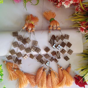 Może przedstawiać: Para ręcznie robionych kolczyków z kwadratowymi, brązowymi koralikami i srebrnymi akcentami. Każdy kolczyk ma pomarańczowe frędzle na dole i na górze. Unikalny dodatek.