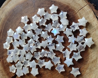 May include: A collection of small, star-shaped gemstones. The stones are a translucent white with iridescent flashes of color. The stars are scattered on a wooden surface, showcasing their faceted surfaces and celestial form. These gemstones are ideal for jewelry making.