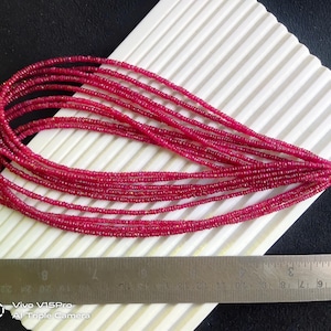 May include: A strand of deep red ruby beads, strung together in a long, continuous line. The beads are small and uniform in size, creating a smooth and elegant look. The strand is laid out on a white surface, with a ruler visible in the foreground.