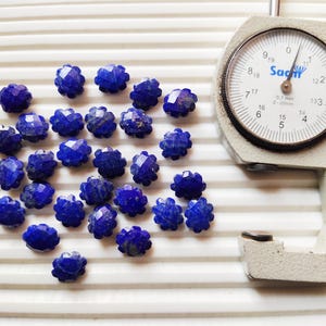 Può includere: Una collezione di perline di pietre preziose blu scuro sfaccettate, simili a piccoli fiori, sono sparse su una superficie bianca. Un misuratore a quadrante argentato, che misura in millimetri, è posizionato a destra.
