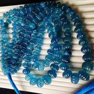 Pode incluir: Um fio de contas de pedras preciosas azuis, cada conta é facetada e tem uma textura única, ligeiramente irregular. As contas são enfiadas em um cordão azul com borlas em cada extremidade.