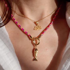 Puede incluir: Un conjunto de collares superpuestos que incluye una cadena dorada con un colgante de flor de loto y un cordón multicolor en rosa, amarillo y azul con un dije de pez dorado sujeto a un cierre de anillo de metal.