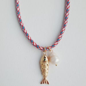 Puede incluir: Un collar con un cordón trenzado en rojo, blanco y azul. El cordón sostiene dos dijes dorados: un colgante de pez texturizado con un pequeño detalle de ojo verde y una perla blanca irregular que cuelga junto al dije de pez.