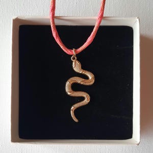 Puede incluir: Un colgante de serpiente de metal en tono dorado suspendido de un cordón trenzado de color coral con un fino hilo metálico dorado. El colgante presenta una superficie texturizada y escamada, presentado en una caja blanca con interior de terciopelo negro.