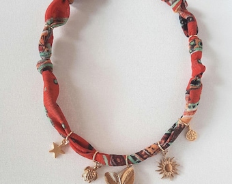 May include: A necklace featuring a red fabric band with a multi-colored pattern, secured by a gold-toned clasp. The band is adorned with several gold-colored metal charms, including a star, a turtle, a large flower, a sun, and a small circular disc.