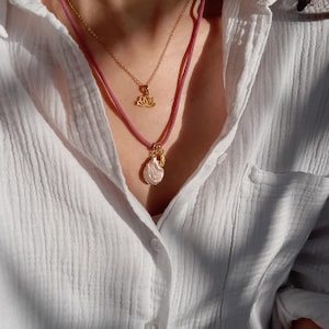 May include: Two necklaces with gold pendants. One features a lotus flower, the other a shell-shaped pendant. The necklaces are layered, with a pink cord necklace and a gold chain. The background is a white button-down shirt.