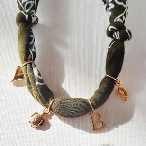 Könnte beinhalten: Halskette aus olivgrünem Stoff mit weißem Paisley-Muster und goldfarbenen Anhängern. Die Anhänger umfassen eine Schildkröte, eine Glocke, ein Blatt und den Buchstaben "B". Die Halskette ist oben geknotet.