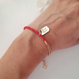 Puede incluir: Una pulsera de cordón rojo con una placa de metal dorado grabada con el texto Good Luck. La pulsera incluye un segmento de cadena dorada con un pequeño dije en forma de trébol en el extremo, lo que permite un ajuste regulable.
