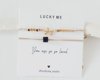Può includere: Due braccialetti delicati su un cartoncino bianco con la scritta LUCKY ME e You are so so loved. Il braccialetto superiore presenta perline metalliche dorate e un ciondolo a forma di foglia. Quello inferiore ha una perlina quadrata blu.