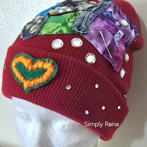 May include: A red knit beanie with a heart-shaped patch and decorative rhinestones. The hat features patches of colorful fabric and the text "Simply Rona".