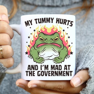 May include: White ceramic mug with a green frog illustration in flames. The text on the mug reads "My tummy hurts and I'm mad at the government."