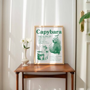 May include: A framed green and white poster with the word "Capybara" in large green letters. The poster also includes a drawing of a capybara, a small bird, and a bucket. The text on the poster reads "(kap-uh-bahr-uh)" and "World's Largest Rodent".