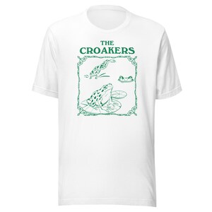 May include: White t-shirt with a green graphic of three frogs on lily pads. The graphic is framed with a decorative border and the text "THE CROAKERS" is above the frogs.