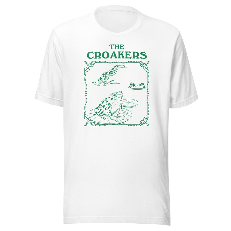 May include: White t-shirt with a green graphic of three frogs on lily pads. The graphic is framed with a decorative border and the text "THE CROAKERS" is above the frogs.