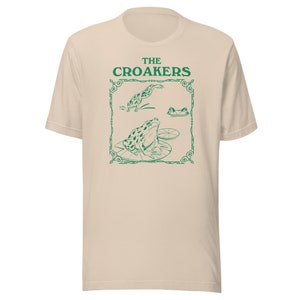 May include: A beige t-shirt with a green graphic of frogs on lily pads. The graphic is framed with a decorative border and the text "THE CROAKERS" is above the frogs.