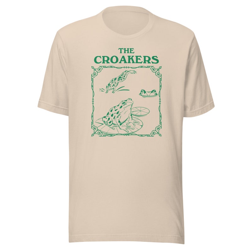 May include: A beige t-shirt with a green graphic of frogs on lily pads. The graphic is framed with a decorative border and the text "THE CROAKERS" is above the frogs.