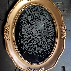 May include: An oval-shaped gold-coloured frame with ornate floral detailing at the top and bottom edges. The frame surrounds a black background featuring a white, intricate spider web design that radiates from a central point towards the edges.
