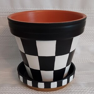 May include: A decorative terracotta flower pot with a black and white checkered pattern. The rim and the matching saucer are also black and white checkered. The interior of the pot is orange. The pot is designed for indoor or outdoor use.