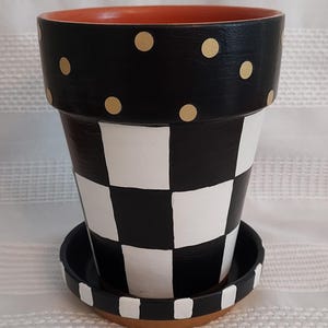 May include: A decorative ceramic flower pot with a black and white checkered pattern on the body. The rim is black with gold polka dots. The pot sits on a matching saucer. The interior is orange.